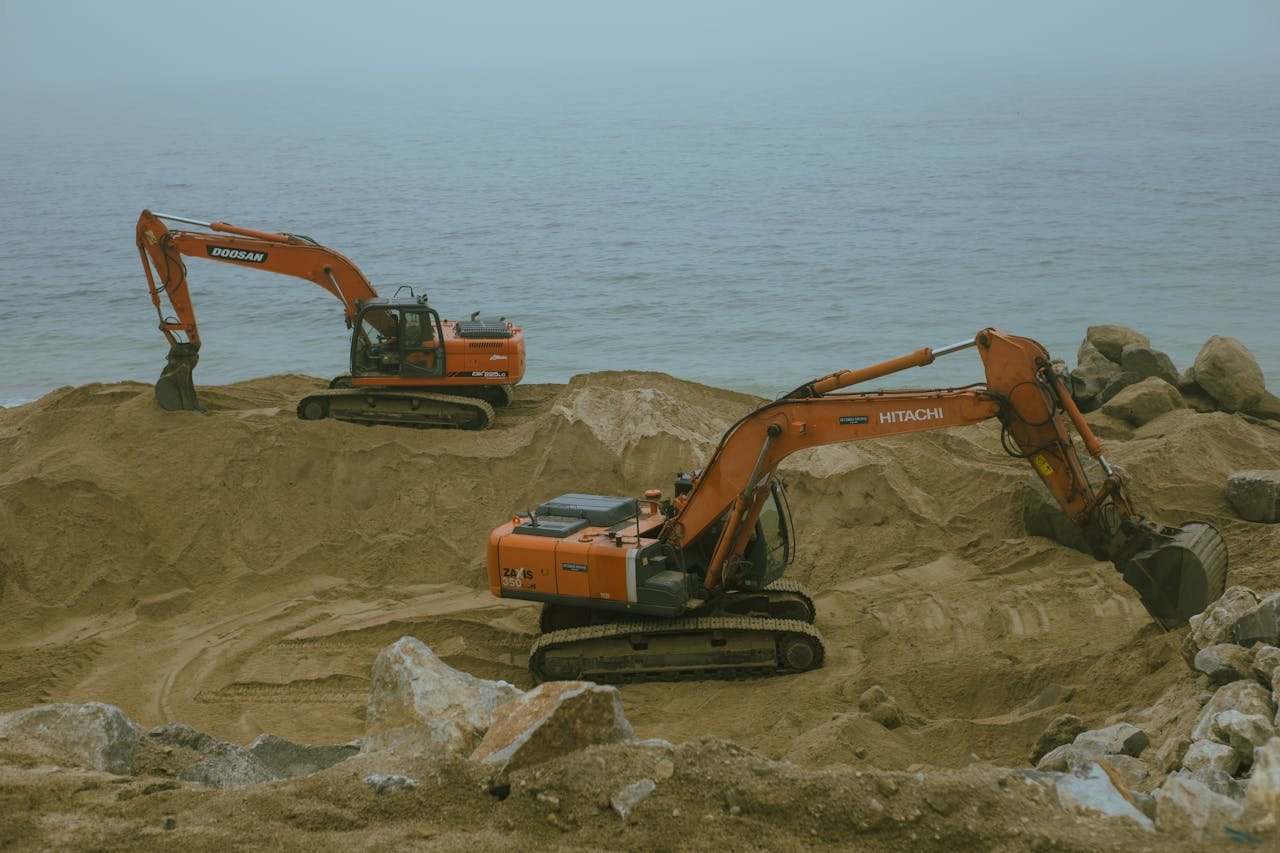 Two excavators operate at a sandy construction site by the sea, moving sand and rock.