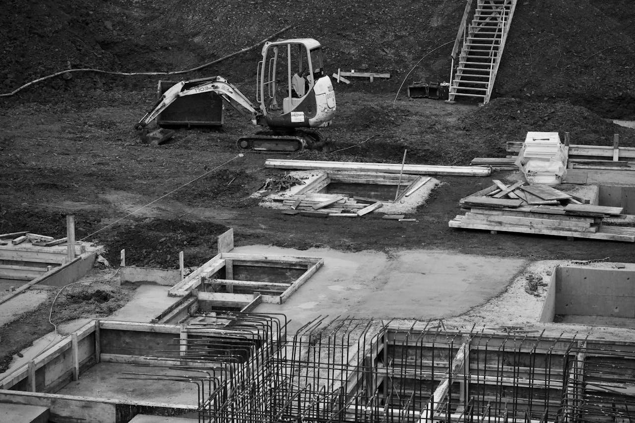 about-me Monochrome image of an excavator at a construction site with foundations laid and rebar in place.